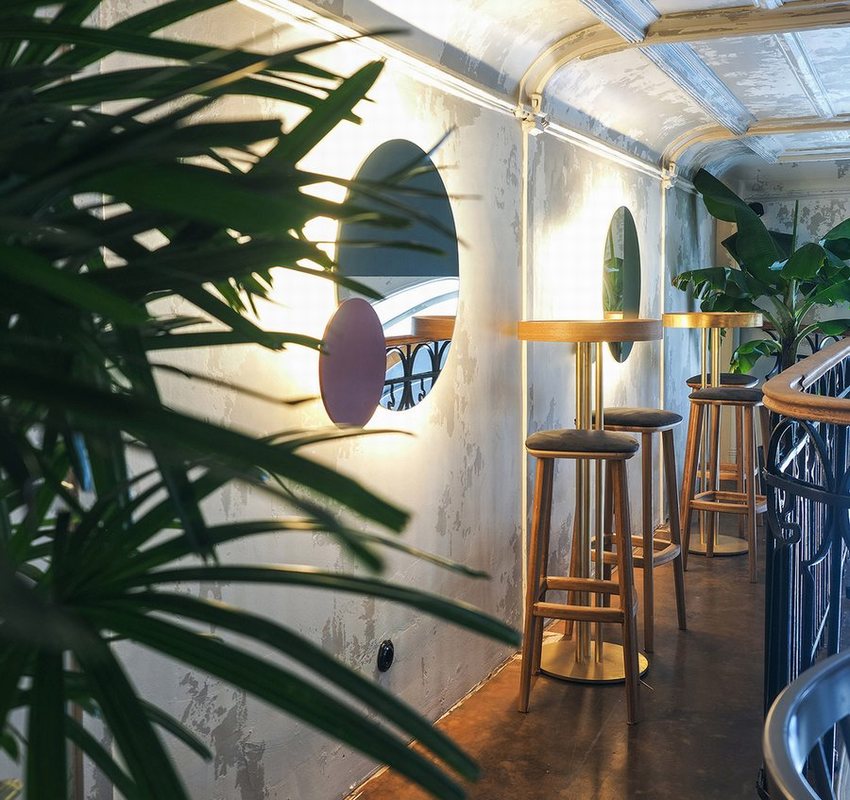 Không gian cafe bar tầng lửng với bàn tròn cao và ghế gỗ tối giản. Tường bê tông thô kết hợp gương tròn nghệ thuật và cây xanh, tạo điểm nhấn độc đáo, mang lại cảm giác vừa hiện đại vừa thư giãn.