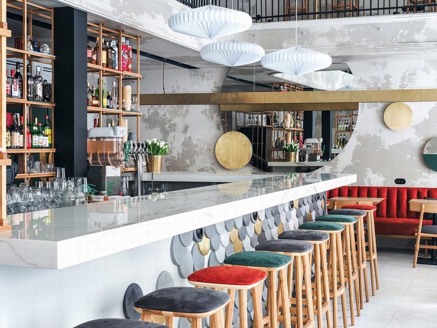 Không gian cafe bar sang trọng với quầy bar mặt đá trắng, ốp họa tiết vảy cá độc đáo. Ghế gỗ bọc nệm nhiều màu sắc kết hợp sofa đỏ nổi bật, cùng hệ thống kệ rượu gỗ và đèn treo nghệ thuật tạo nên vẻ hiện đại, thu hút.
