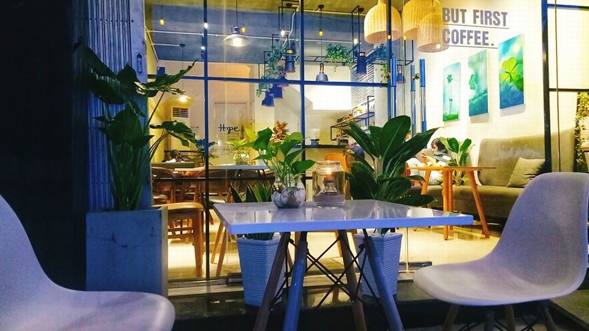 Không gian quán cafe hiện đại kết hợp cây xanh trang trí, bàn ghế tối giản và ánh sáng ấm áp, tạo cảm giác gần gũi và thư giãn.