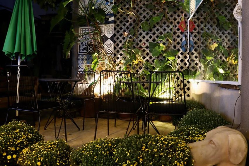 Không gian quán cafe sân vườn buổi tối với ánh sáng trang trí ấm áp, bàn ghế ngoài trời và cây xanh leo tường tạo điểm nhấn tự nhiên.