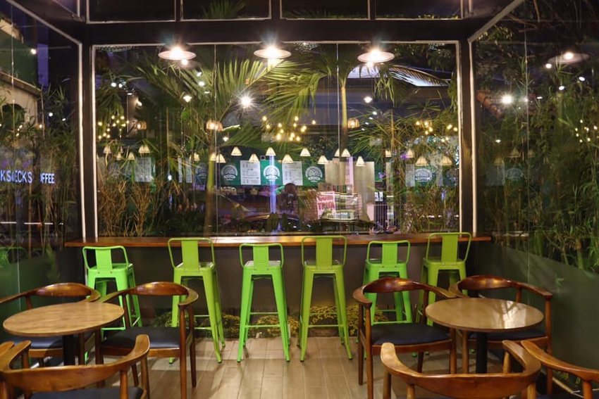 Thiết kế nội thất quán cafe sân vườn với không gian bên trong ấm cúng, bàn ghế gỗ kết hợp ghế cao hiện đại, view kính nhìn ra cây xanh thoáng đãng.