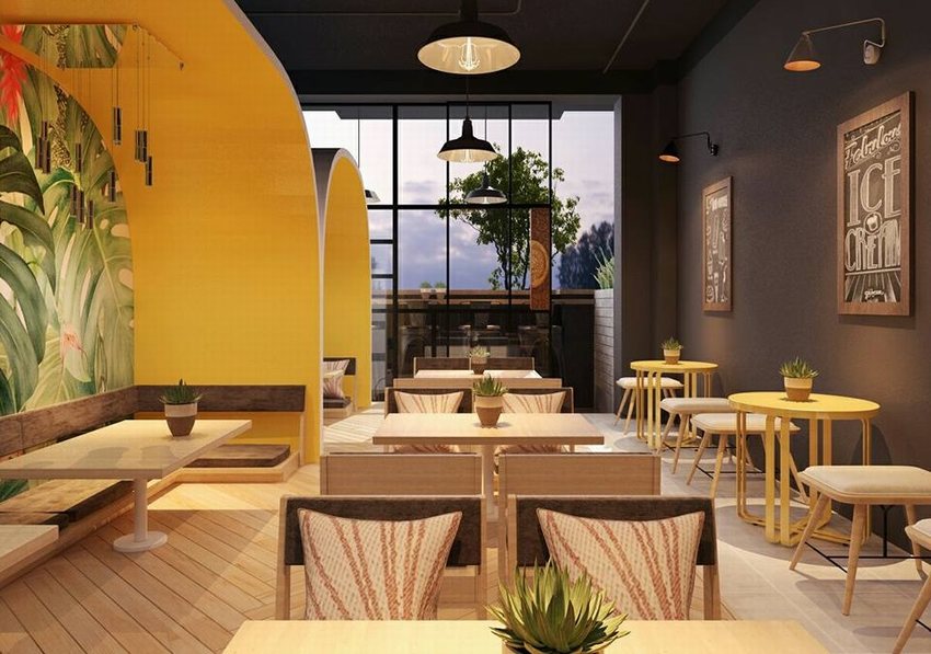 Không gian quán cafe trà sữa tươi sáng với tường vàng, bàn ghế gỗ, tranh tường lá cây nhiệt đới, cửa kính lớn và cây xanh tạo cảm giác gần gũi thiên nhiên.