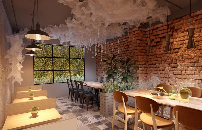 Khu vực ngồi của quán cafe trà sữa với tường gạch đỏ, bàn gỗ dài, trần trang trí vải trắng buông rủ, ánh sáng vàng ấm và cửa kính lớn hướng ra vườn cây xanh.