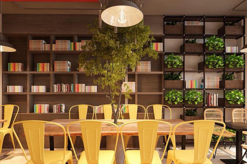 Không gian quán cafe với kệ sách lớn chứa đầy sách, cây xanh trang trí, ghế sắt màu vàng, bàn gỗ dài và ánh sáng vàng ấm áp, mang phong cách thư viện hiện đại.