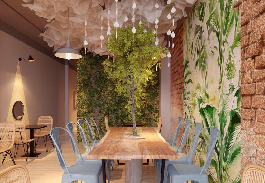 Quán cafe trà sữa với bàn dài, ghế kim loại xanh, cây xanh ở giữa bàn, tường trang trí tranh lá cây, gạch đỏ và trần vải trắng buông rủ cùng đèn thả tạo không gian thư giãn ấm cúng.