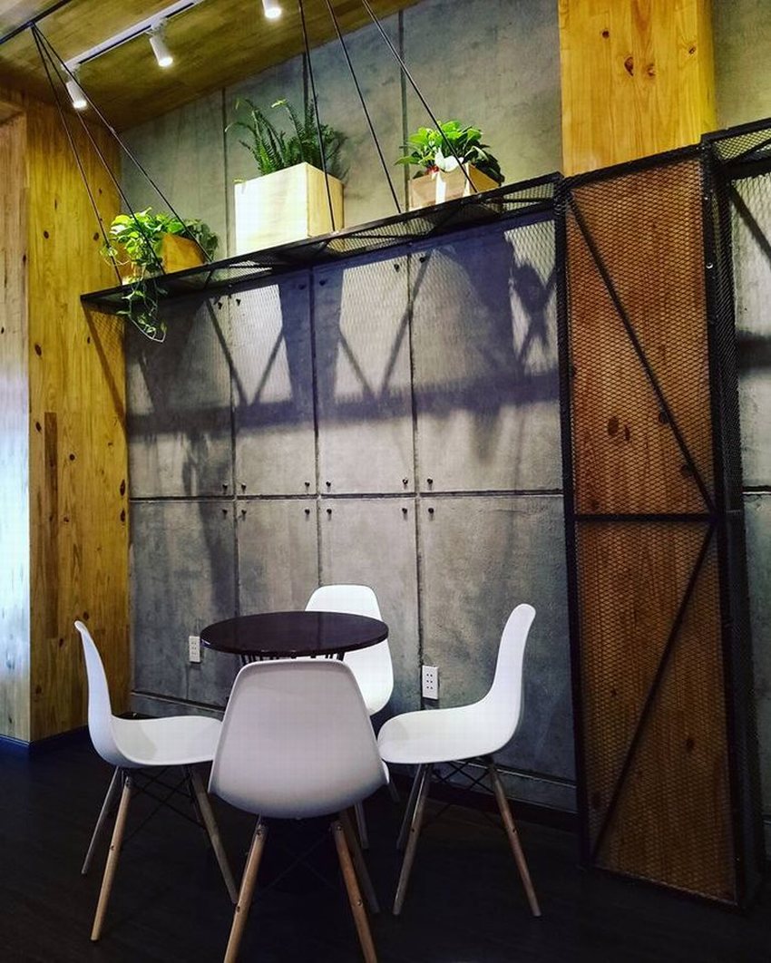 Góc ngồi nhỏ trong quán cafe trà sữa, bàn tròn gỗ với ghế nhựa trắng chân gỗ, tường bê tông thô kết hợp gỗ ấm áp, trang trí cây xanh treo trên kệ sắt.