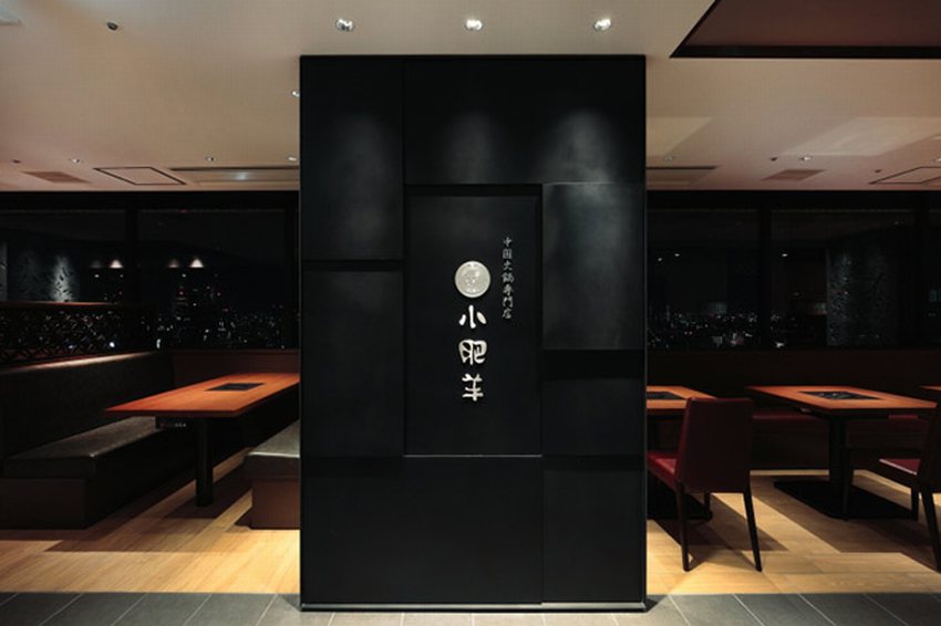 Bức tường trung tâm với logo nhà hàng được thiết kế hiện đại, sử dụng chất liệu đen nhám sang trọng, nổi bật với dòng chữ và biểu tượng khắc nổi theo phong cách Nhật Bản. Phía sau là không gian bàn ăn ấm cúng với tầm nhìn thành phố về đêm, tạo nên sự kết hợp hài hòa giữa thương hiệu và trải nghiệm không gian.