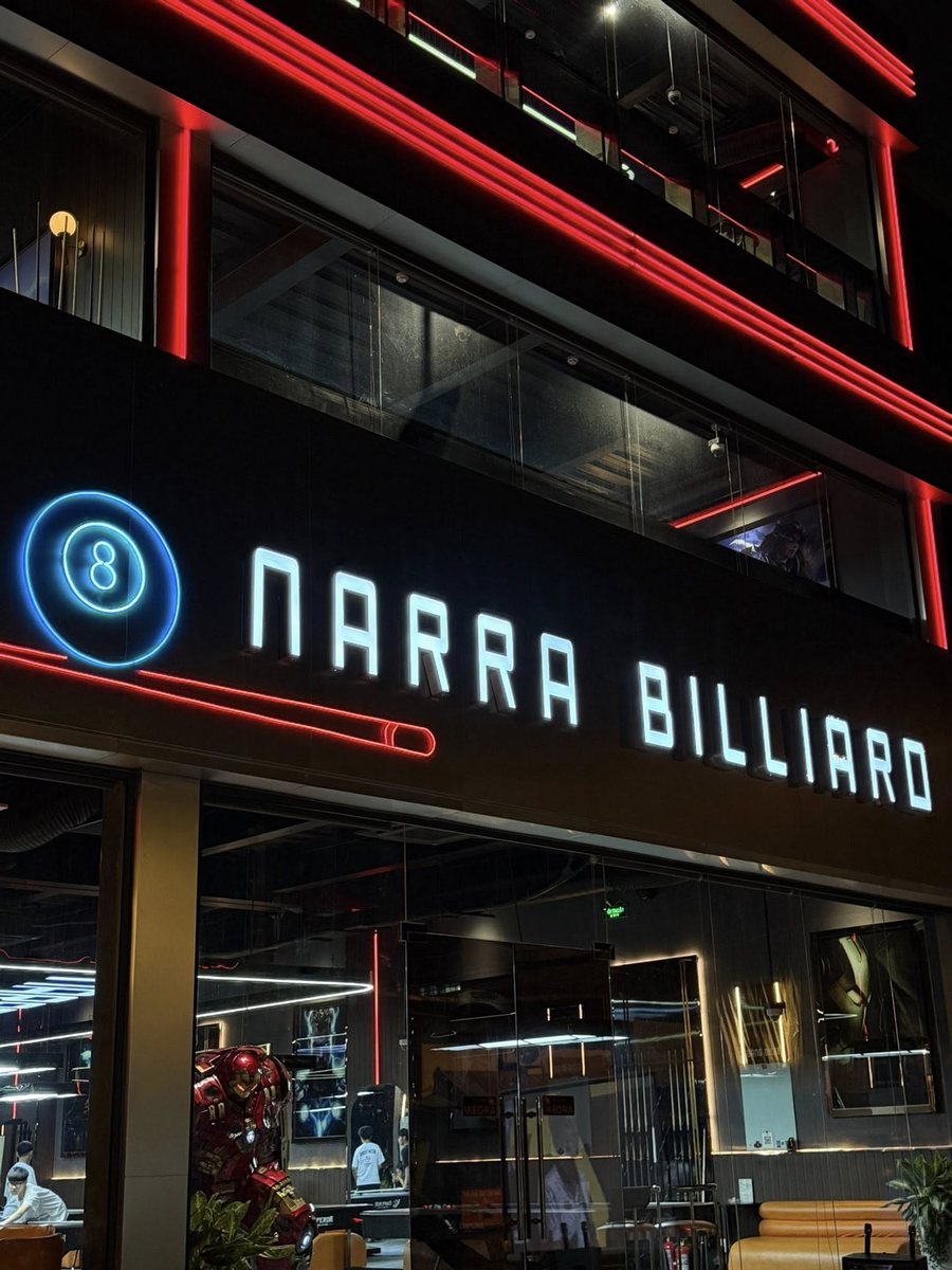 Mặt tiền NARRA BILLIARDS gây ấn tượng mạnh với thiết kế hiện đại, cá tính. Biển hiệu chữ nổi phát sáng kết hợp biểu tượng bi số 8 tạo điểm nhận diện rõ ràng, dễ ghi nhớ. Hệ đèn LED đỏ – xanh neon chạy viền kiến trúc làm nổi khối công trình về đêm, mang hơi hướng Dark Luxury – Industrial. Không gian kính lớn mở tầm nhìn vào bên trong, vừa sang trọng vừa năng động, thu hút khách ngay từ ánh nhìn đầu tiên. 