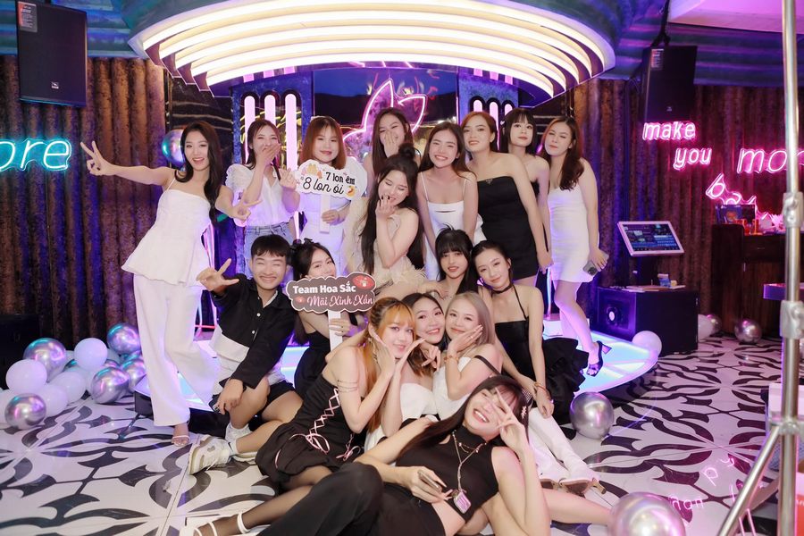 Khu vực lounge bar với ánh sáng neon, sân khấu nhỏ và nhóm khách đông vui chụp ảnh kỷ niệm, thể hiện rõ góc kinh doanh đẹp kết hợp tổ chức sự kiện, giúp tăng trải nghiệm khách hàng và thu hút nhóm khách trẻ năng động